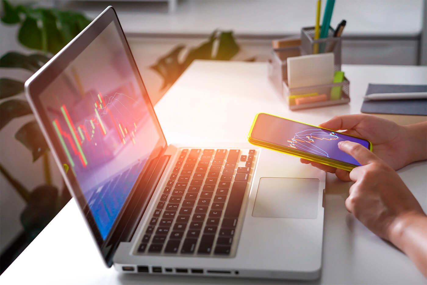 A hand holds a phone above a computer screen displaying financial graphs, possibly exploring Financial Solutions with the help of Continentalink.