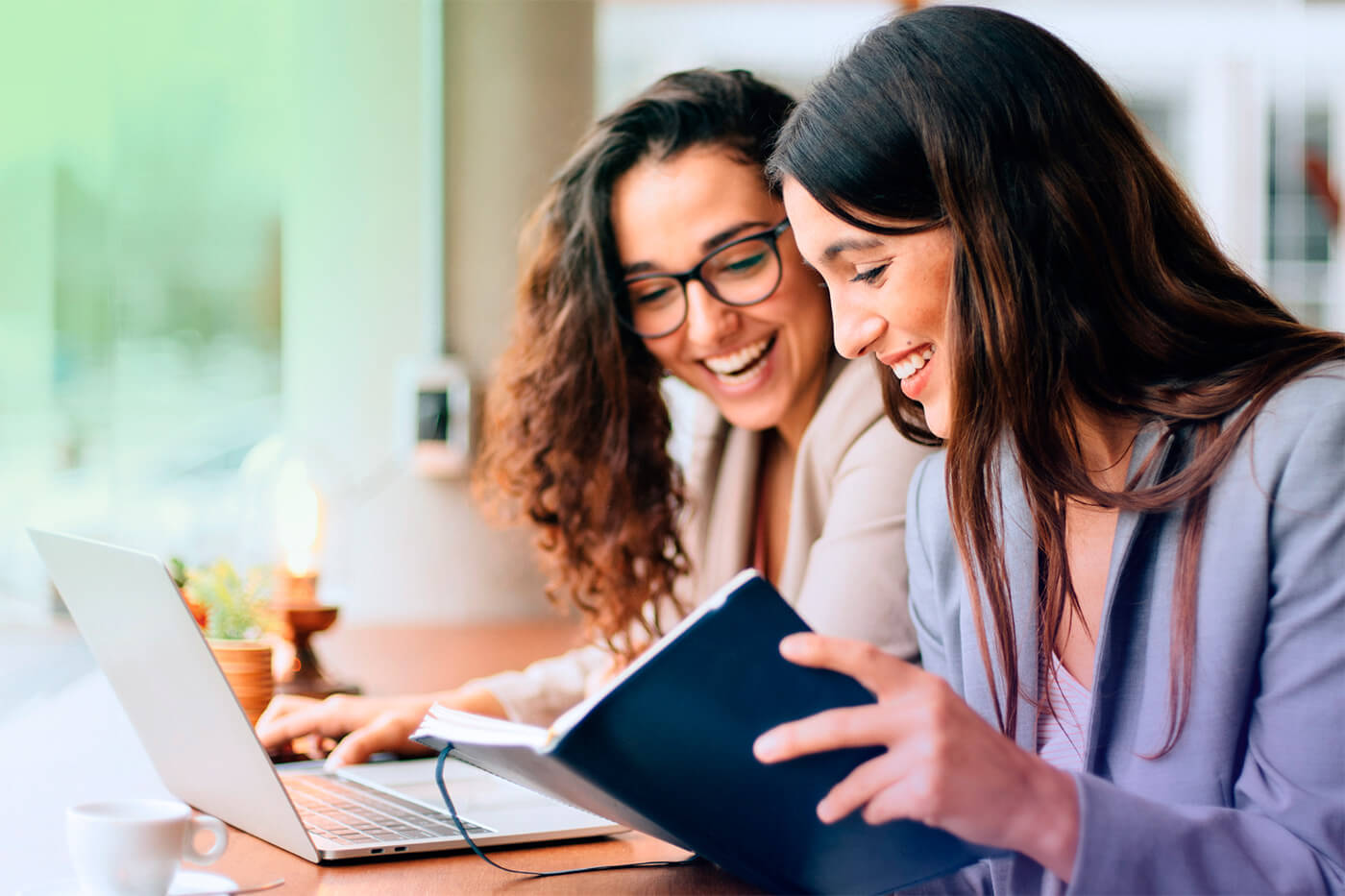 Two women seated at a desk, sharing laughter while looking at a computer screen and notebook, possibly discussing Financial Solutions and Continentalink.